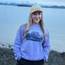Person wearing a purple sweatshirt with a graphic design, standing by a body of water with mountains in the background.