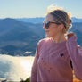 A woman wearing a dusty pink long-sleeve crewneck sweatshirt with a small black heart and star graphic, standing outdoors on a hillside with a scenic mountain and ocean view in the background.