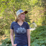 A woman wearing a navy blue v-neck t-shirt with 'TREETOP TEES' printed on the front, blue jeans, and a trucker hat, standing outdoors in a lush green forest.