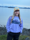 Person wearing a purple sweatshirt with a graphic design, standing by a body of water with mountains in the background.