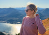 A woman wearing a dusty pink long-sleeve crewneck sweatshirt with a small black heart and star graphic, standing outdoors on a hillside with a scenic mountain and ocean view in the background.