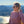 A woman wearing a dusty pink long-sleeve crewneck sweatshirt with a small black heart and star graphic, standing outdoors on a hillside with a scenic mountain and ocean view in the background.