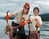 A woman and a child standing in a boat on an overcast day, each holding a large red crab. Both are wearing matching white graphic T-shirts, orange gloves, and winter hats.
