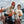 A woman and a child standing in a boat on an overcast day, each holding a large red crab. Both are wearing matching white graphic T-shirts, orange gloves, and winter hats.