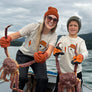A woman and a child standing in a boat on an overcast day, each holding a large red crab. Both are wearing matching white graphic T-shirts, orange gloves, and winter hats.