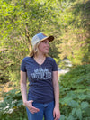 A woman wearing a navy blue v-neck t-shirt with 'TREETOP TEES' printed on the front, blue jeans, and a trucker hat, standing outdoors in a lush green forest.